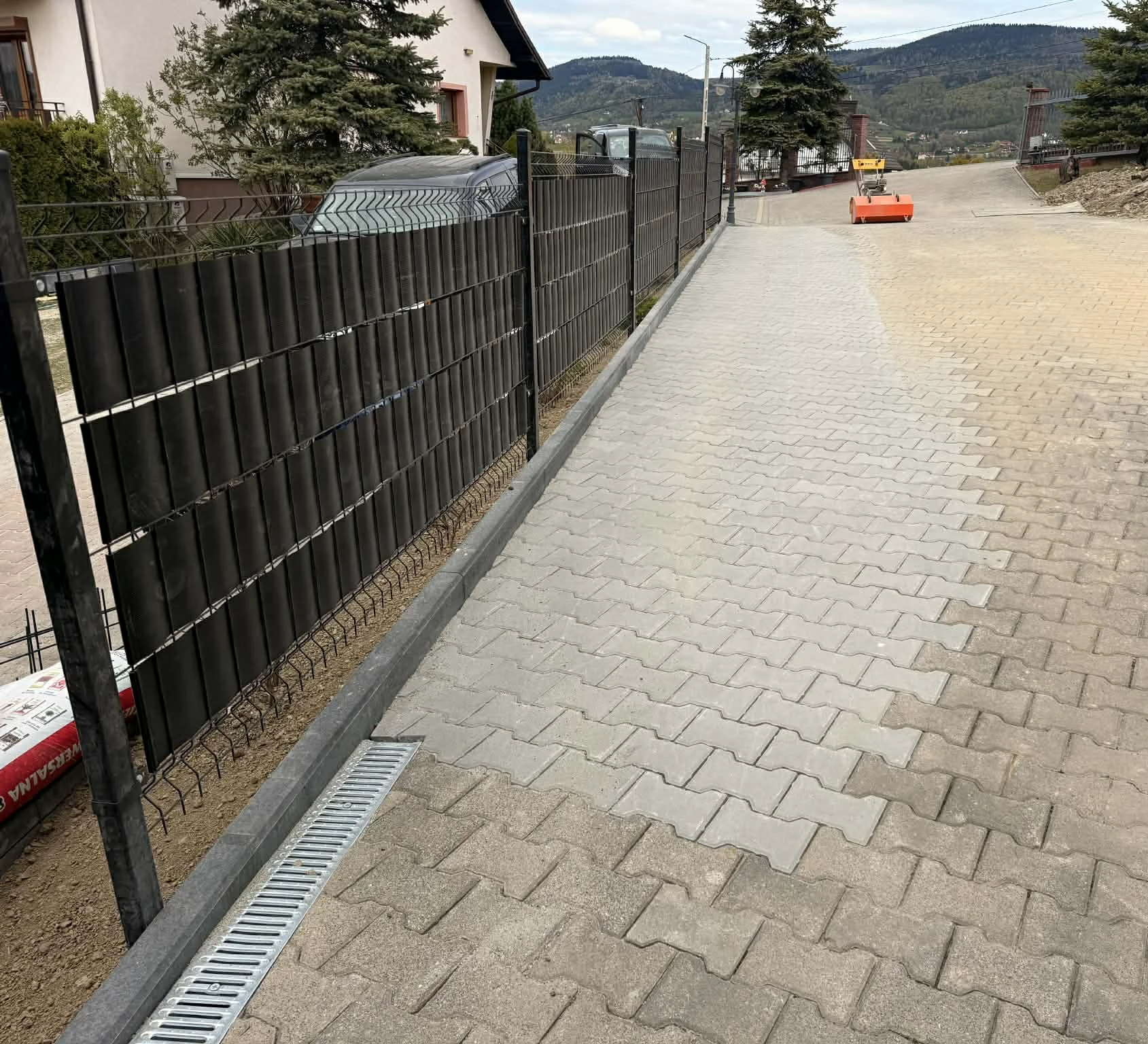 Układanie kostki brukowej: nowy chodnik i podjazd z szarej kostki betonowej przy ogrodzeniu panelowym z linią odpływu